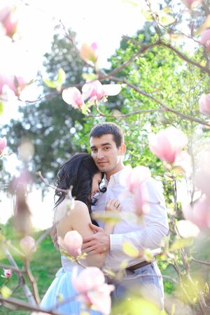 Brunette handsome boy hugging and kissing caucasian girl near magnolia blooming tree. Concept of love and spring season.の写真素材