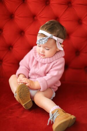 Little caucasian girl wearing pink sweater sitting on sofa with red background. Concept of baby photo session.の写真素材