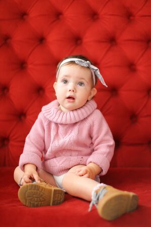 Little nice girl wearing pink sweater sitting on sofa with red background. Concept of baby photo session.の写真素材
