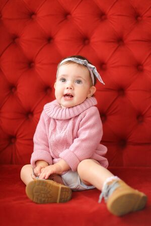 Little smiling girl wearing pink sweater sitting on sofa with red background. Concept of baby photo session.の写真素材