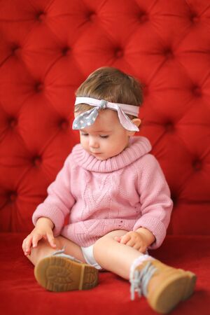 Little happy girl wearing pink sweater sitting on sofa with red background. Concept of baby photo session.の写真素材