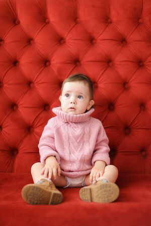 Little beautiful girl wearing pink sweater sitting on sofa with red background. Concept of baby photo session.の写真素材