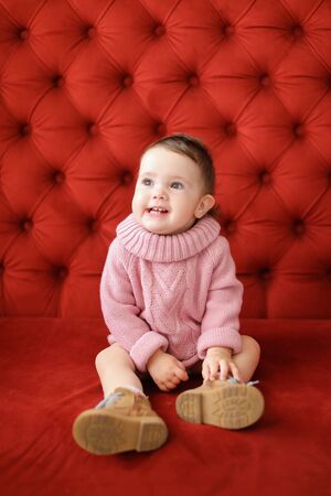 Little cute female baby sitting in red background on sofa. Concept of babias and photo session.の写真素材