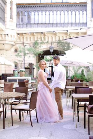 Happy caucasian groom and fiancee standing at restaurant near table and building in background. Concept of gladden young couple and wedding.の写真素材