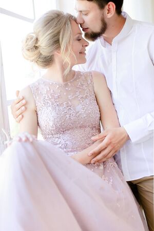 Blonde caucasian bride wearing dress and standing near window with groom. Concept of bridal photo session and nice couple.の写真素材