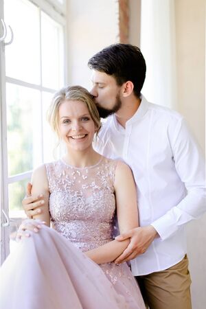 Blonde cute bride wearing dress and standing near window and kissing groom. Concept of bridal photo session and nice couple.の写真素材