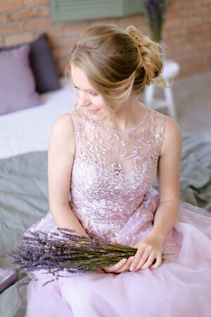 Blonde fiancee sitting with lavander bouquet at home. Concept of bridal photo session in provans way.の写真素材