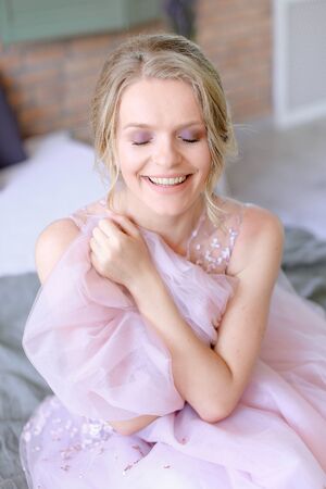 Happy beautiful blonde bride sitting in room and smiling. Concept of bridal photo session and posotive emotions.の写真素材