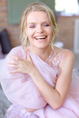 Happy charming blonde bride sitting in room and smiling. Concept of bridal photo session and posotive emotions.の写真素材