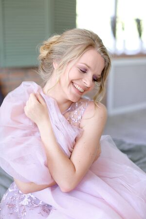 Happy european blonde bride sitting in room and smiling. Concept of bridal photo session and posotive emotions.の写真素材