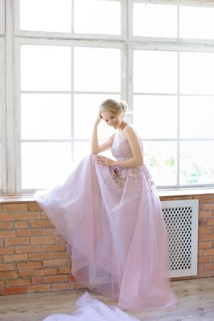 Blonde bride wearing dress and standing near window and dreaming. Concept of bridal photo session and nice fiancee.の写真素材