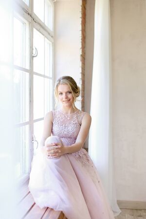 Blonde nice bride wearing dress and standing near window and dreaming. Concept of bridal photo session and nice fiancee.の写真素材