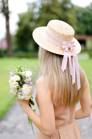 Back view of young blonde girl in body color overalls and hat standing in yeard with flowers. Concept of fashion and summer season.の写真素材