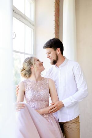 Blonde tender bride wearing dress and standing near window with groom. Concept of bridal photo session and nice couple.の写真素材