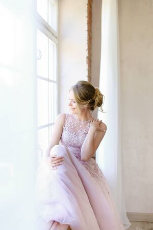 Blonde pretty bride wearing dress and standing near window and dreaming. Concept of bridal photo session and nice fiancee.の写真素材