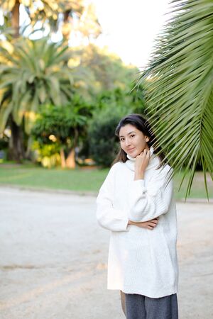 Korean cute woman in white sweater standing near palm leaf. Concept of asian beauty and female person on tropical resort.の写真素材