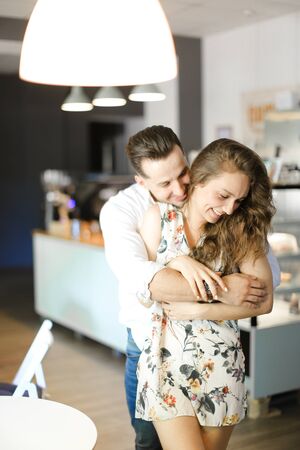 Young smiling man hugging nice girl and standing at cafe near tables. Concept of love and positive feelings.の写真素材