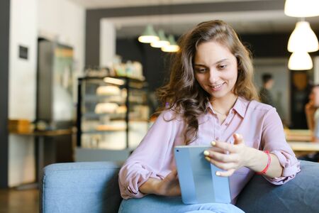 Young female smiling student sitting on sofa at cafe and surfing by tablet. Concept of social networks, modern technology and free time with internet.の写真素材