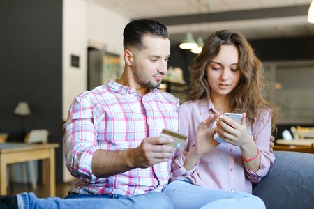 Young caucasian wife and husband using smartphone and debit card at cafe. Concept of purchasing online and shopping, modern technology.の写真素材