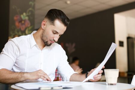Young caucasian handsome businessman working with papers at cafe, wearing white shirt. Concept of business and occupation.の写真素材