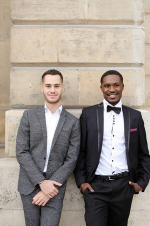 Afro american and caucasian gays standing near building and wearing suits. Concept of lgbt and walking in city.の写真素材