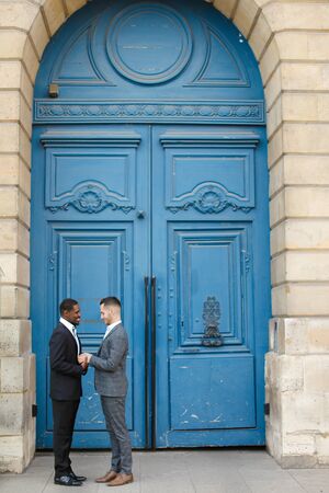 Afro american smiling man holding hands of caucasian boy in blue door background, wearing suit. Concept of same sex couple and gays.の写真素材