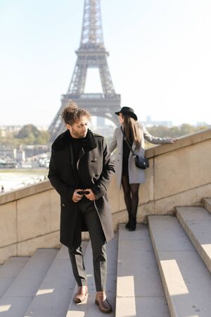Focus on afro american handsome man with curly hair on concrete stairs, girl and Eiffel Tower in background, Paris.の写真素材