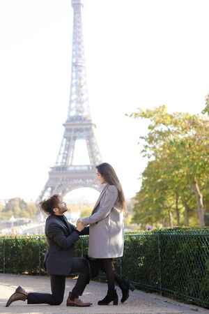 Afro american happy boy making proposal to caucasian girlfriend in Paris, Eiffel Tower in background. Concept of love and international couple.の写真素材