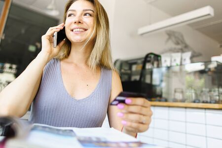 Blonde girl speaking by smartphone and keeping debit card in hands. Concept of shopping online and cheap phone conversation.の写真素材