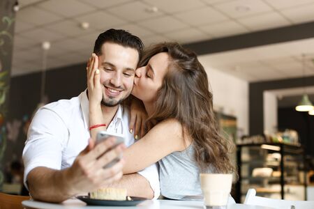 Young wife kissing husband using smartphone at cafe and eating cake. Concept of food and drink, happy couple and relationship, modern technology.の写真素材