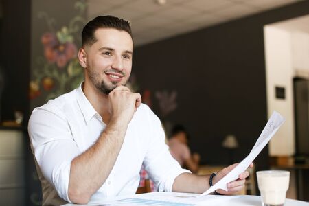 Young happy handsome businessman working with papers at cafe, wearing white shirt. Concept of business and occupation.の写真素材