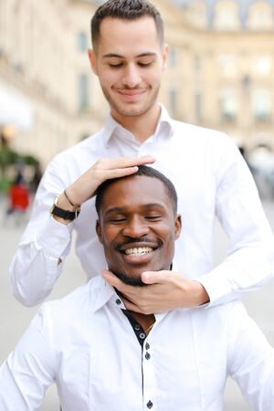 Caucasian boy holding afro american guy head by hands, wearing white shirt. Concept of stylish boy and haircut.の写真素材