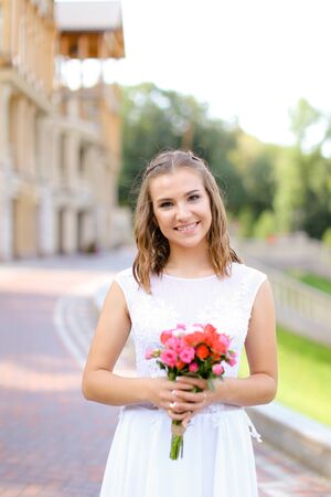 Young bride wearing white dress and standing outside with flowers. Concept of wedding and bridal photo session, floristic art.の写真素材