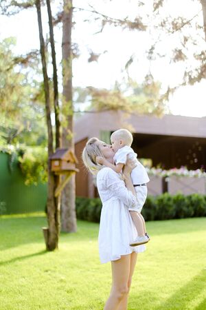 Young blonde mom walking with child on grass in yard. Concept of motherhood and kids, summer season.の写真素材