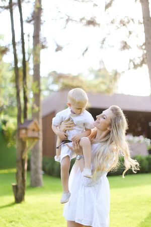 Young caucasian mom walking with child on grass in yard. Concept of motherhood and kids, summer season.の写真素材
