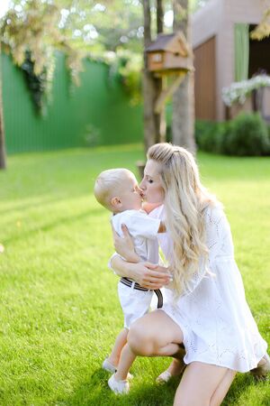 Young happy mother playing and sitting with little kid on grass in yard and kissing son. Concept of motherhood and child.の写真素材