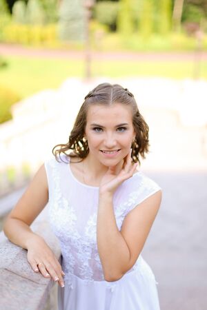 Happy charming bride wearing white dress standing outside and smiling. Concept of bridal photo session and wedding, nice fiancee.の写真素材
