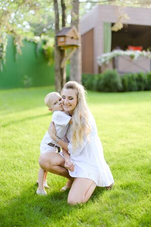 Young happy mother playing and sitting with little kid on grass in yard. Concept of motherhood and child.の写真素材
