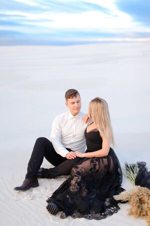 Young girl wearing black dress and man sitting on snow in steppe. Concept of winter photo session and love.の写真素材