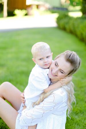Young mom sitting with little child on grass in yard. Concept of motherhood and summer vacations.の写真素材