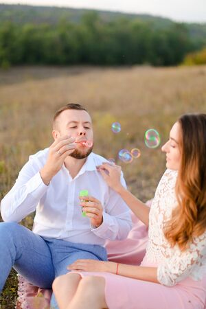 Young boy blowing soap bubbles near caucasian woman and sitting on pink plaid in steppe. Concept of happy couple, romantic love and picnic on nature.の写真素材
