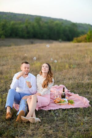Young cheerful boy blowing soap bubbles near caucasian girl and sitting on pink plaid in steppe. Concept of happy couple, romantic love and picnic on nature.の写真素材