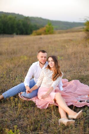 Young beautiful wife and husband sitting on pink plaid in field. Concept of picnic in open air and relationship, love.の写真素材