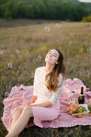 Young pretty woman sitting on plaid with bottle of wine and fruits, soap bubbles. Concept of beauty nad picnic on nature.の写真素材