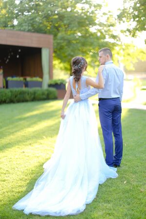 Back view of caucasian fiancee in white dress and and groom walking in garden. oncept of bridal hair do and wedding photo session on nature.の写真素材