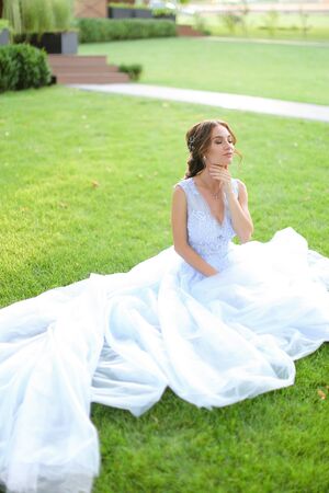 Caucasian pretty bride sitting on grass in park, wearing white amazing dress. Concept of bridal photo session in open air, beautiful fiancee.の写真素材