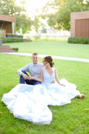 Caucasian bride with groom sitting on grass in park, wearing white amazing dress. Concept of bridal photo session in open air, beautiful fiancee.の写真素材