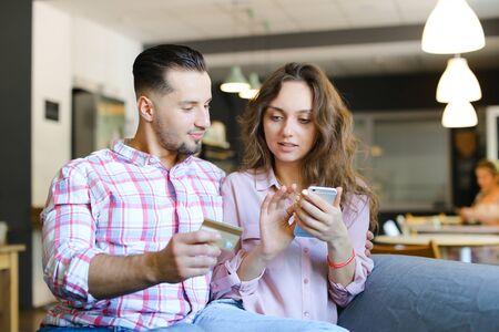 Young cute wife and husband using smartphone and debit card at cafe. Concept of purchasing online and shopping, modern technology.の写真素材