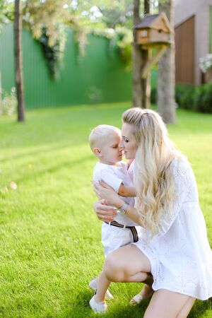 Young caucasian mother playing and sitting with little kid on grass in yard and kissing son. Concept of motherhood and child.の写真素材