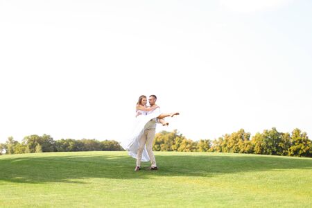 Caucasian groom holding bride on grass in white sky background. Concept of wedding photo session on nature.の写真素材